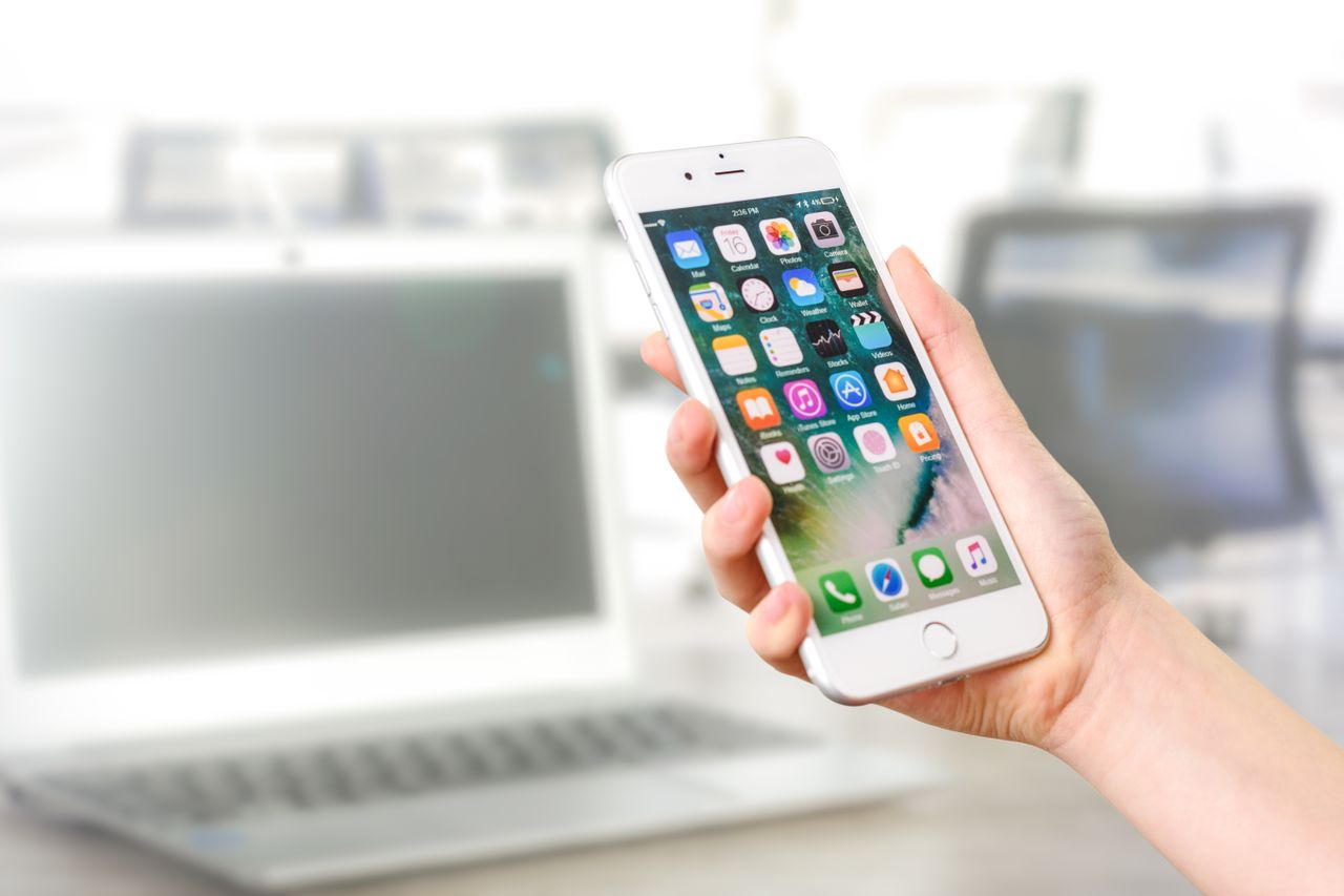 Home Close-up view of a smartphone displaying apps, held by a hand, with a blurred laptop in the background.